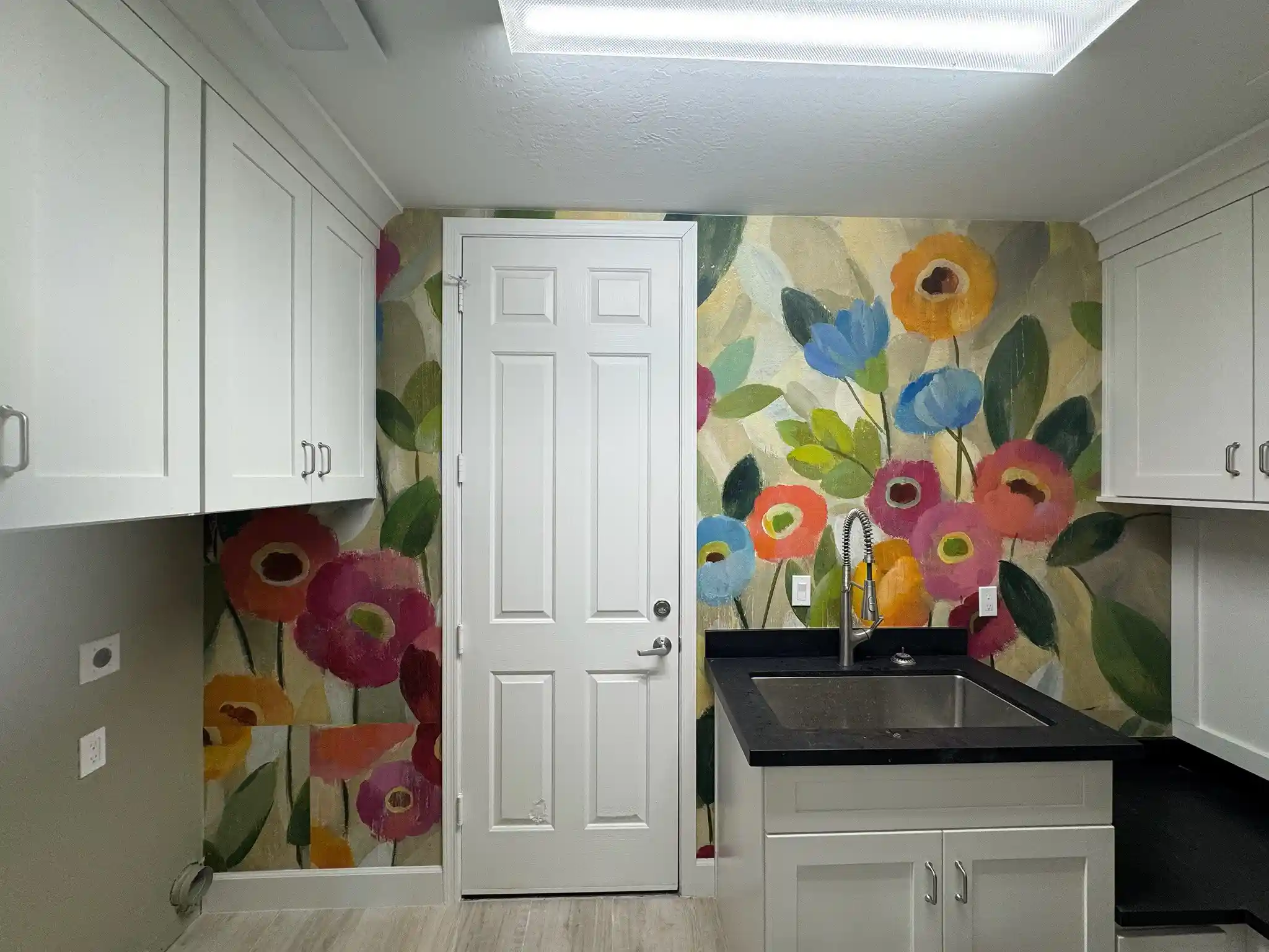 White utility room with cabinetry, a black sink and oversized floral wallcovering behind the door and counter.