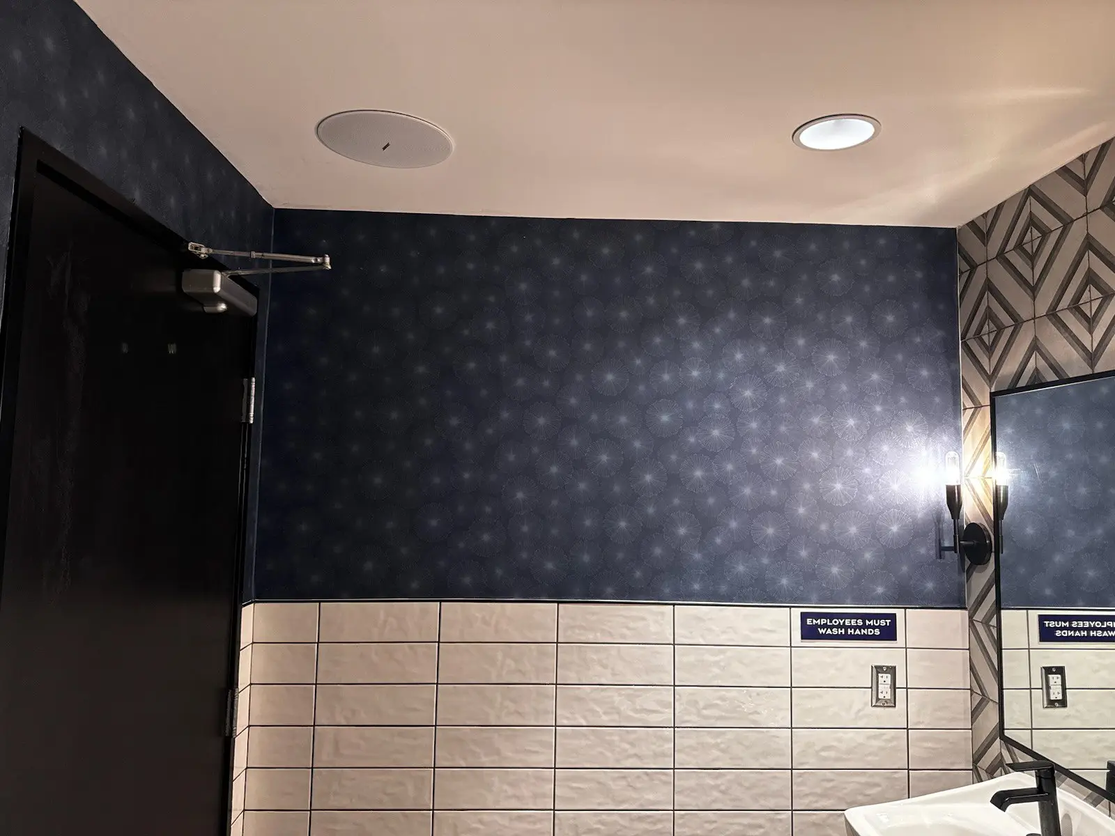 Dark blue radial-pattern wallpaper above white subway tile, beside a black door, mirror and wall sconces.